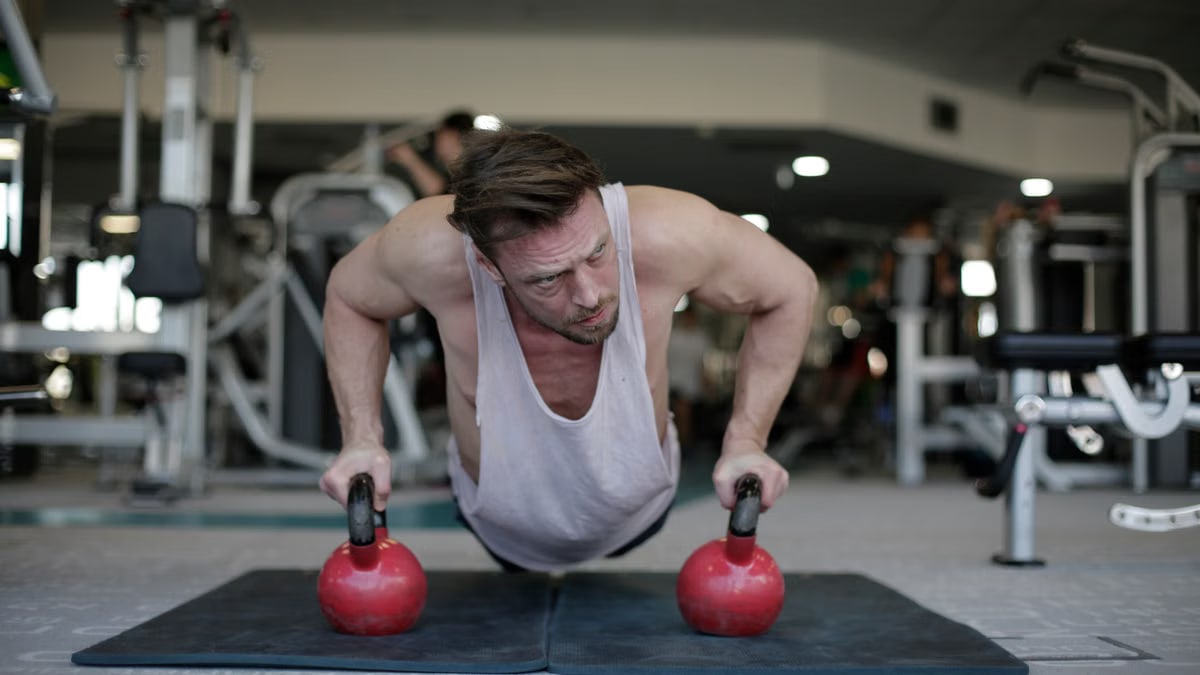 Adult male athlete wearing activewear performing push up exercise with kettlebells during fitness workout on floor of modern spacious sport club and looking away